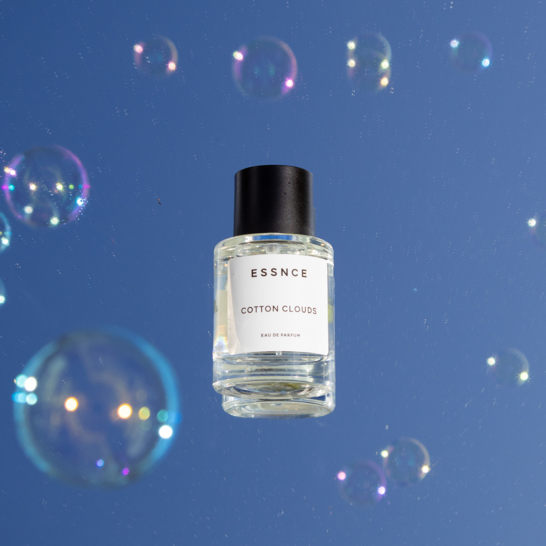 A bottle of ESSNCE Cotton Clouds perfume, featuring an elegant and minimalist design with a black cap, floats against a blue background adorned with colorful, iridescent bubbles.