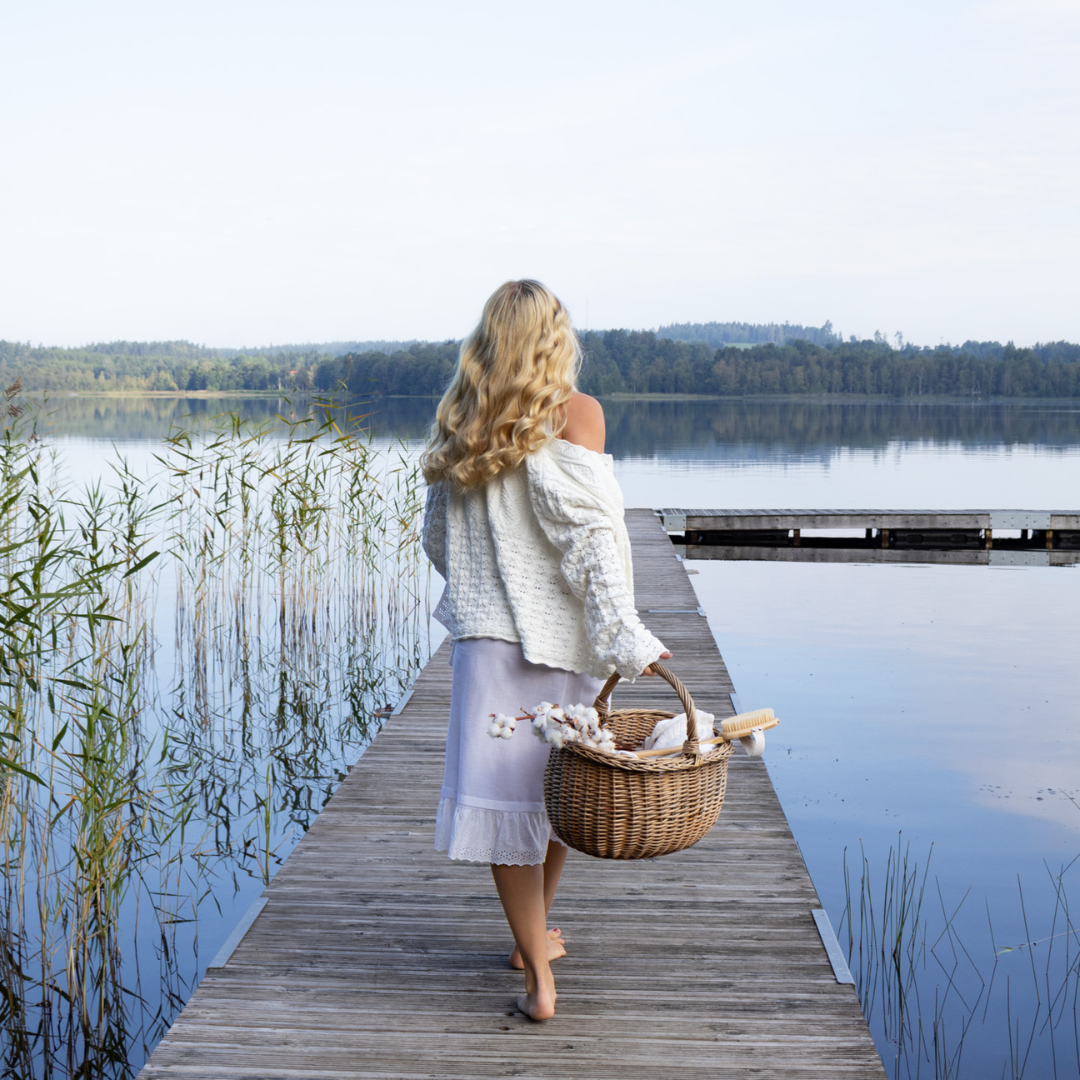 A woman with long blonde hair, dressed in a white sweater and a light purple skirt, walks barefoot on a wooden dock towards a serene lake. She carries a woven basket filled with flowers and bread. The scene is tranquil, with reeds and a forested shoreline in the background.