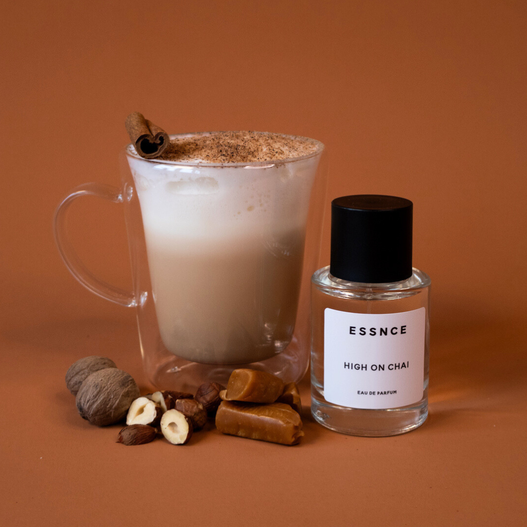 En glaskopp med chai latte omgiven av hasselnötter, karameller, muskotnötter och en stilfull parfymflaska märkt ESSNCE High on Chai Eau de Parfum, vilket skapar en elegant och inbjudande scen.